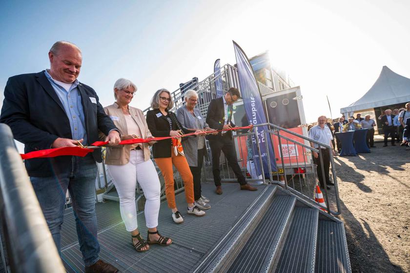 EN: Five people cut a red ribbon to open the viewing platform. They are standing on the steps leading up to the viewing platform, which is behind them. // DE: Fünf Personen schneiden ein rotes Band zur Eröffnung des neuen Aussichtspunkts durch. Sie stehen auf der Treppe zur Aussichtsplattform, die sich hinter den Personen befindet. // DA: Fem personer klipper et rødt bånd over ved åbningen af udsigtsplatformen. De står på trappen til udsigtsplatformen, som befinder sig bag personerne.
