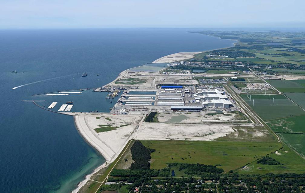 Das Übersichtsfoto zeigt in Form eines Luftbilder die Baustelle auf Lolland aus Osten mit der Küstenlinie, dem Arbeitshafen und der Fabrik für die Tunnelelemente.