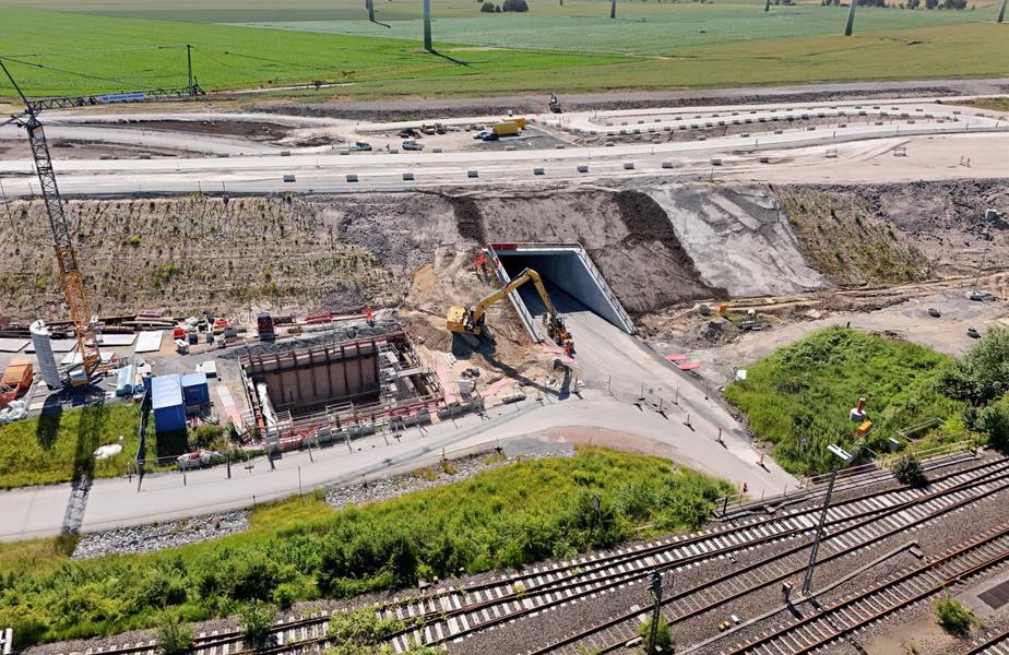 Das Bild zeigt einen großen Damm, auf dem später die Autobahn E47 verlaufen wird. Eine Unterführung unter diesem Damm ist zu sehen, durch die der neue Weg nach Marienleuchte in Kürze geführt wird. Links daneben ist eine große Baugrube mit Baugerät, Arbeitern und einem Kran zu sehen. Unten im Bild sind die Gleise der alten Bahntrasse.