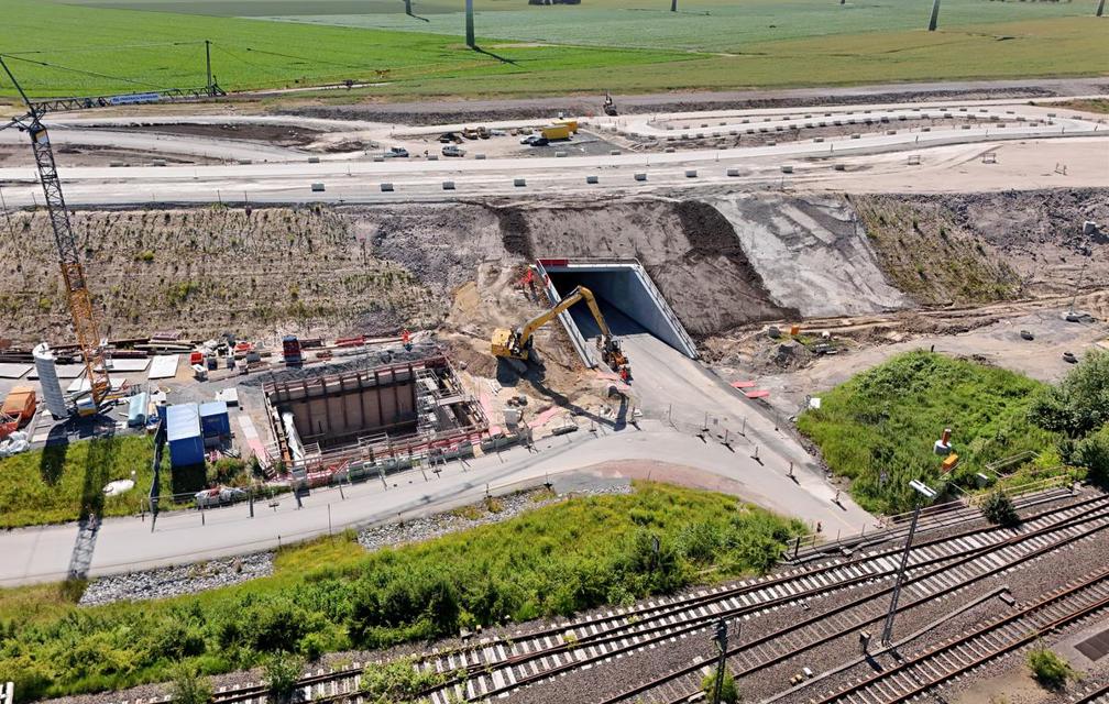 Das Bild zeigt einen großen Damm, auf dem später die Autobahn E47 verlaufen wird. Eine Unterführung unter diesem Damm ist zu sehen, durch die der neue Weg nach Marienleuchte in Kürze geführt wird. Links daneben ist eine große Baugrube mit Baugerät, Arbeitern und einem Kran zu sehen. Unten im Bild sind die Gleise der alten Bahntrasse.