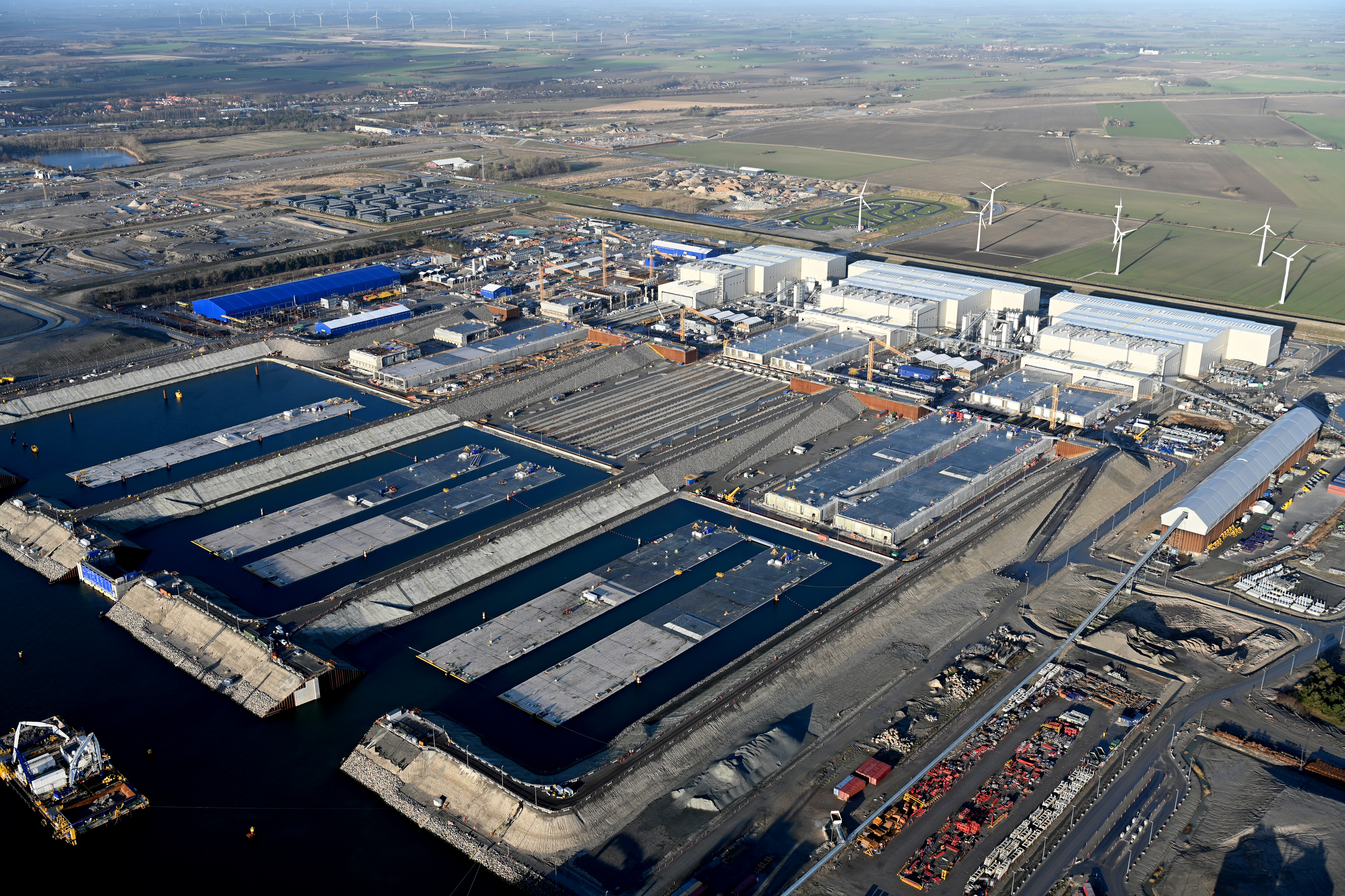 Tunnelelementfabrikken ved Rødbyhavn. Man kan se de tre store hvide fabrikshaller, hvor tunnelelementerne fremstilles. Foran fabrikshallerne ligger færdigstøbte elementer. Nogle flyder allerede i vandet.