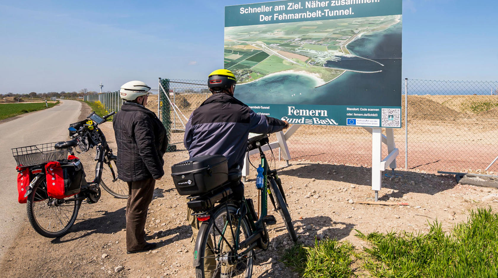 EN: Two cyclists standing in front of an information sign next to the construction site. // DE: Zwei Fahrradfahrer stehen vor einem Informationsschild neben der Baustelle. // DA: To cyklister står foran et informationsskilt ved siden af byggepladsen.