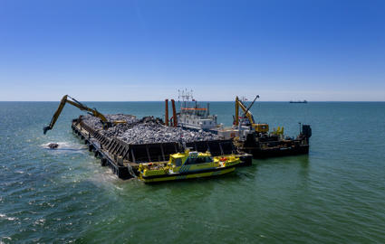 English: A barge with granit for the breakwaters in the Fehmarnbelt. Excavators drop the boulders into the sea to construct the breakwaters.//Dansk: En pram med granit til bølgebrydere i Fehmarnbelt. Gravemaskiner lægger kampestenene i havet for at bygge bølgebryderne.//Deutsch: Eine Schute mit Granit für die Wellenbrecher im Fehmarnbelt. Bagger versenken die Granitbrocken im Meer, um die Wellenbrecher zu errichten.