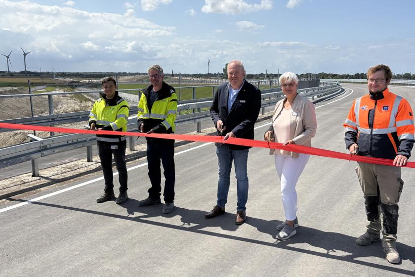 EN: Five people cut a red ribbon to open the Marienleuchter Weg. The new road can be seen. // DE: Fünf Personen schneiden ein rotes Band zur Eröffnung der Marienleuchter Wegs durch. Man sieht die neue Straße. // DA: Fem personer klipper et rødt bånd over ved åbningen af Marienleuchter Weg. Man kan se den nye vej.
