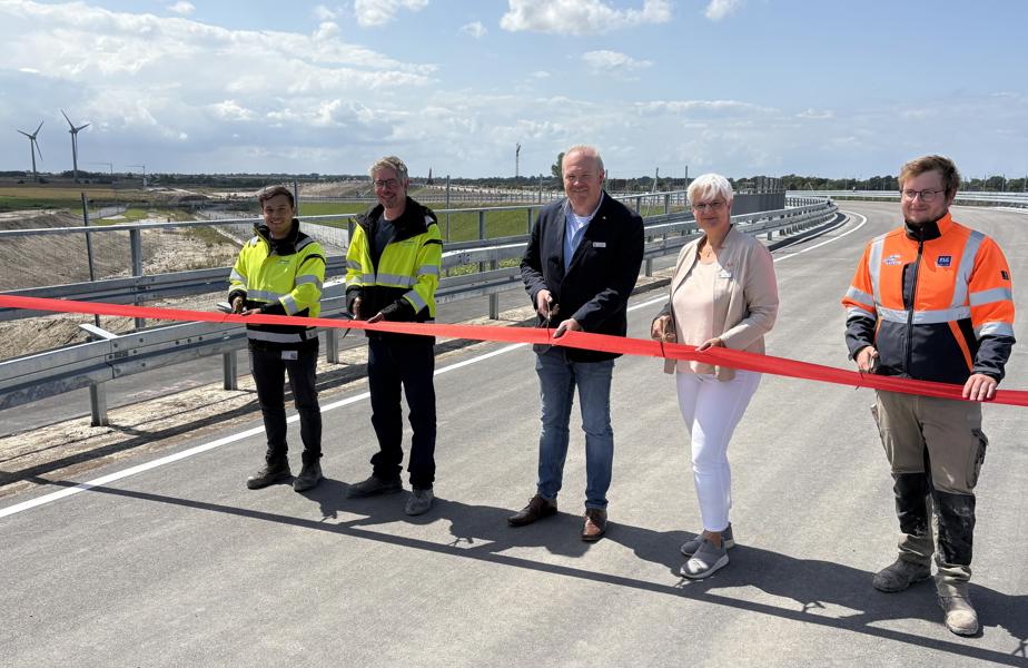 EN: Five people cut a red ribbon to open the Marienleuchter Weg. The new road can be seen. // DE: Fünf Personen schneiden ein rotes Band zur Eröffnung der Marienleuchter Wegs durch. Man sieht die neue Straße. // DA: Fem personer klipper et rødt bånd over ved åbningen af Marienleuchter Weg. Man kan se den nye vej.