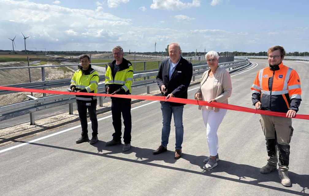EN: Five people cut a red ribbon to open the Marienleuchter Weg. The new road can be seen. // DE: Fünf Personen schneiden ein rotes Band zur Eröffnung der Marienleuchter Wegs durch. Man sieht die neue Straße. // DA: Fem personer klipper et rødt bånd over ved åbningen af Marienleuchter Weg. Man kan se den nye vej.