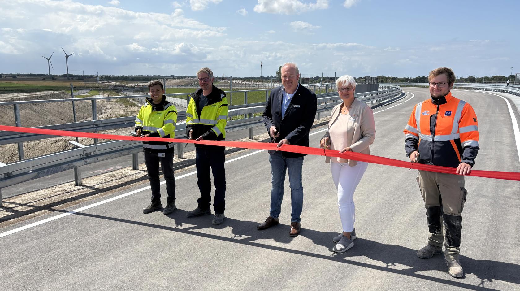 EN: Five people cut a red ribbon to open the Marienleuchter Weg. The new road can be seen. // DE: Fünf Personen schneiden ein rotes Band zur Eröffnung der Marienleuchter Wegs durch. Man sieht die neue Straße. // DA: Fem personer klipper et rødt bånd over ved åbningen af Marienleuchter Weg. Man kan se den nye vej.