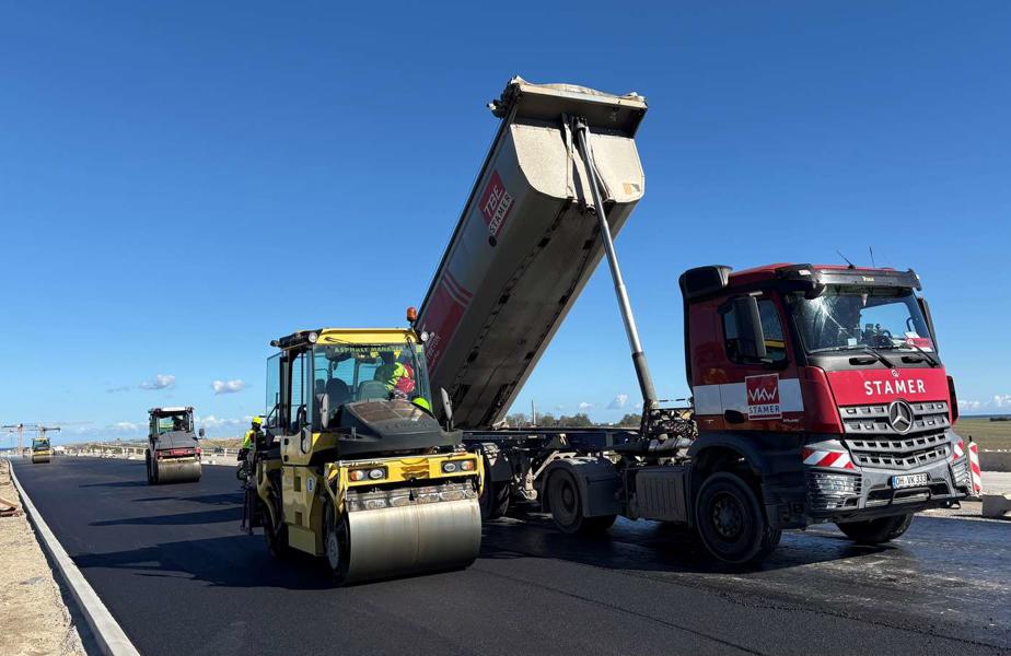 Asphaltierungsarbeiten auf der neuen Straßenanbindung, ein Lkw und eine Straßenwalze bringen den Fahrbahnbelag auf, weitere Baumaschinen arbeiten im Hintergrund.