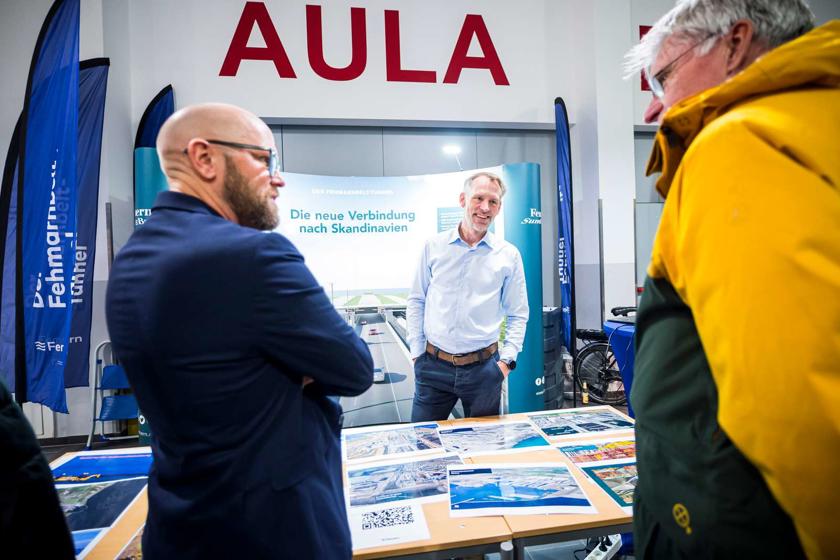 Drei Männer stehen um einen Tisch, auf dem Bilder von der Tunnelbaustelle liegen. Einer der Männer ist ein Bürger, die beiden Experten von Femern A/S beantworten seine Fragen.