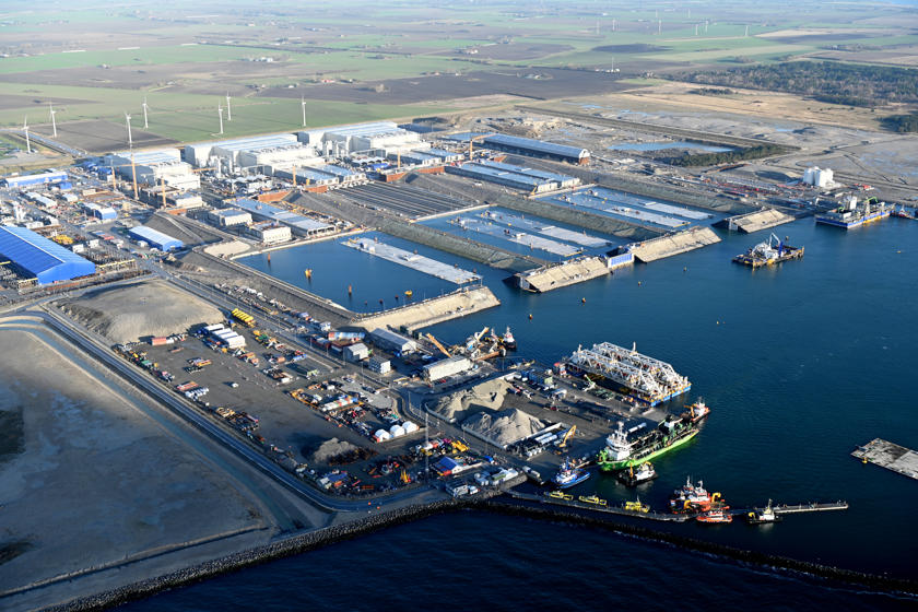 The tunnel factory on Lolland. Large concrete tunnel elements are produced in three large white halls. In front of the halls are water basins through which the elements are floated. Some elements float in the water.