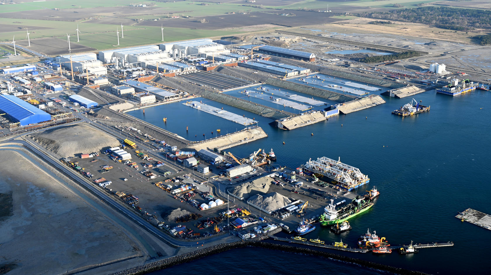 The tunnel factory on Lolland. Large concrete tunnel elements are produced in three large white halls. In front of the halls are water basins through which the elements are floated. Some elements float in the water.
