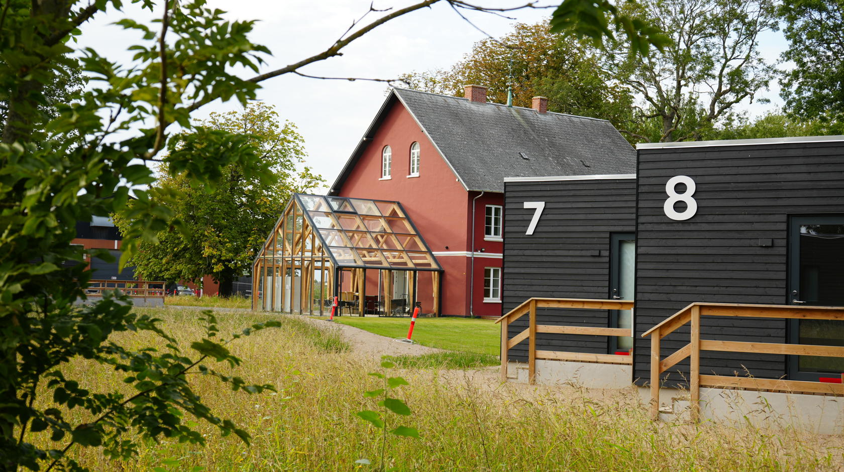 The red main building of Campus Femern with an attached conservatory. In the foreground are two residential huts with wooden fronts.