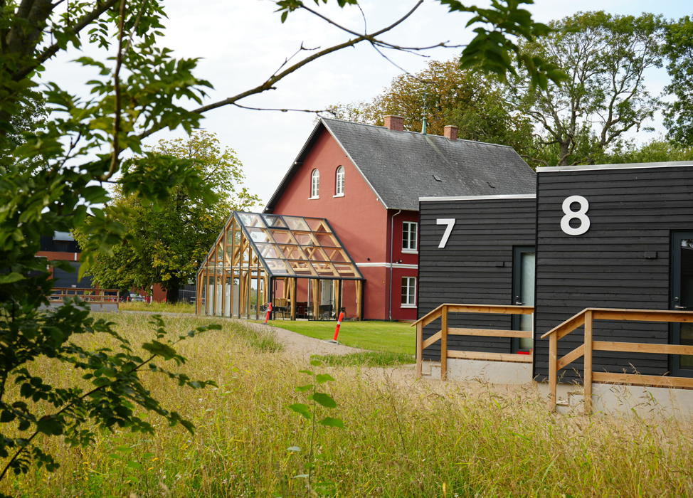 Das rote Haupthaus vom Campus Femern mit angebauten Wintergarten. Im Vordergrund zwei Wohnhütten mit Holzfassade.
