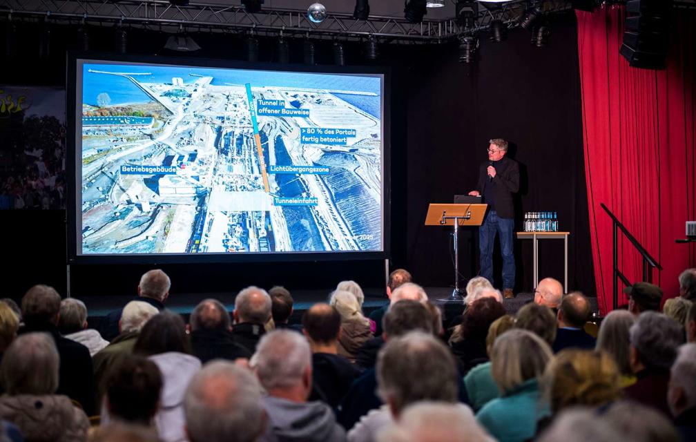 Gregory Formichella steht auf einer Bühne und hält einen Vortrag über den Fehmarnbelt-Tunnel. Auf der Leinwand neben ihm sieht man ein Bild von der Baustelle. Man sieht die Hinterköpfe vieler Menschen im Publikum.
