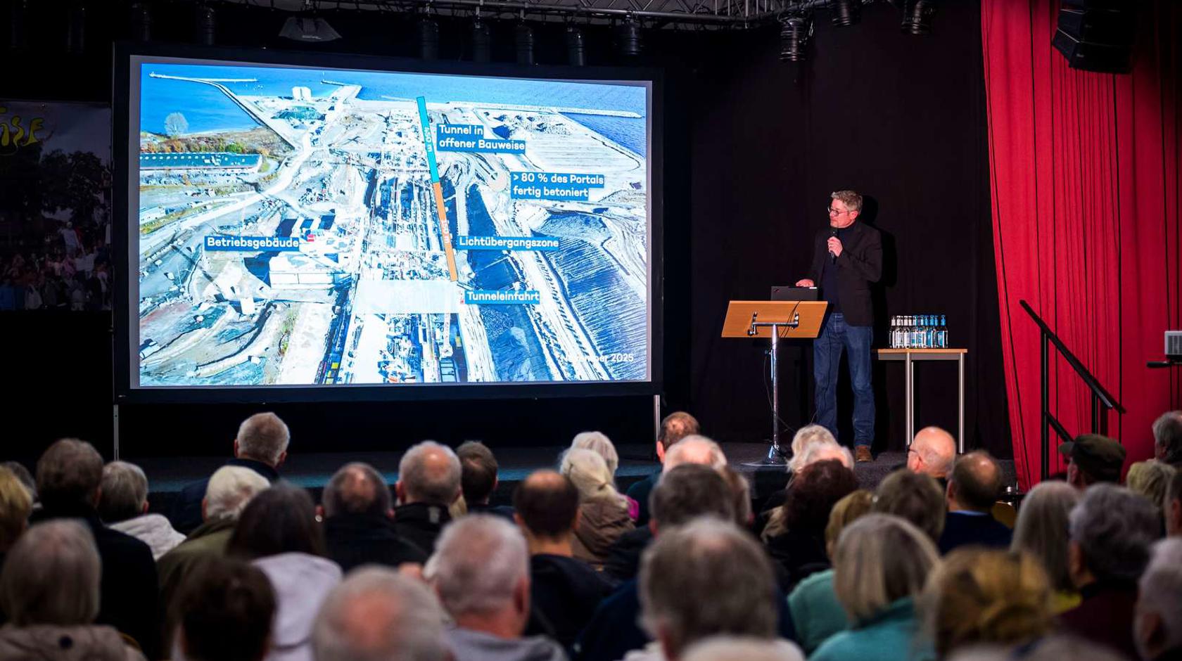 Gregory Formichella steht auf einer Bühne und hält einen Vortrag über den Fehmarnbelt-Tunnel. Auf der Leinwand neben ihm sieht man ein Bild von der Baustelle. Man sieht die Hinterköpfe vieler Menschen im Publikum.