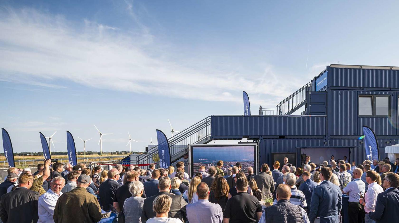 EN: Around 200 people stand in front of a large screen and listen to a speech. // DE: Rund 200 Personen stehen vor einem großen Bildschirm und hören einer Rede zu. // DK: Rundt om 200 personer står foran en stor skærm og lytter til en tale.
