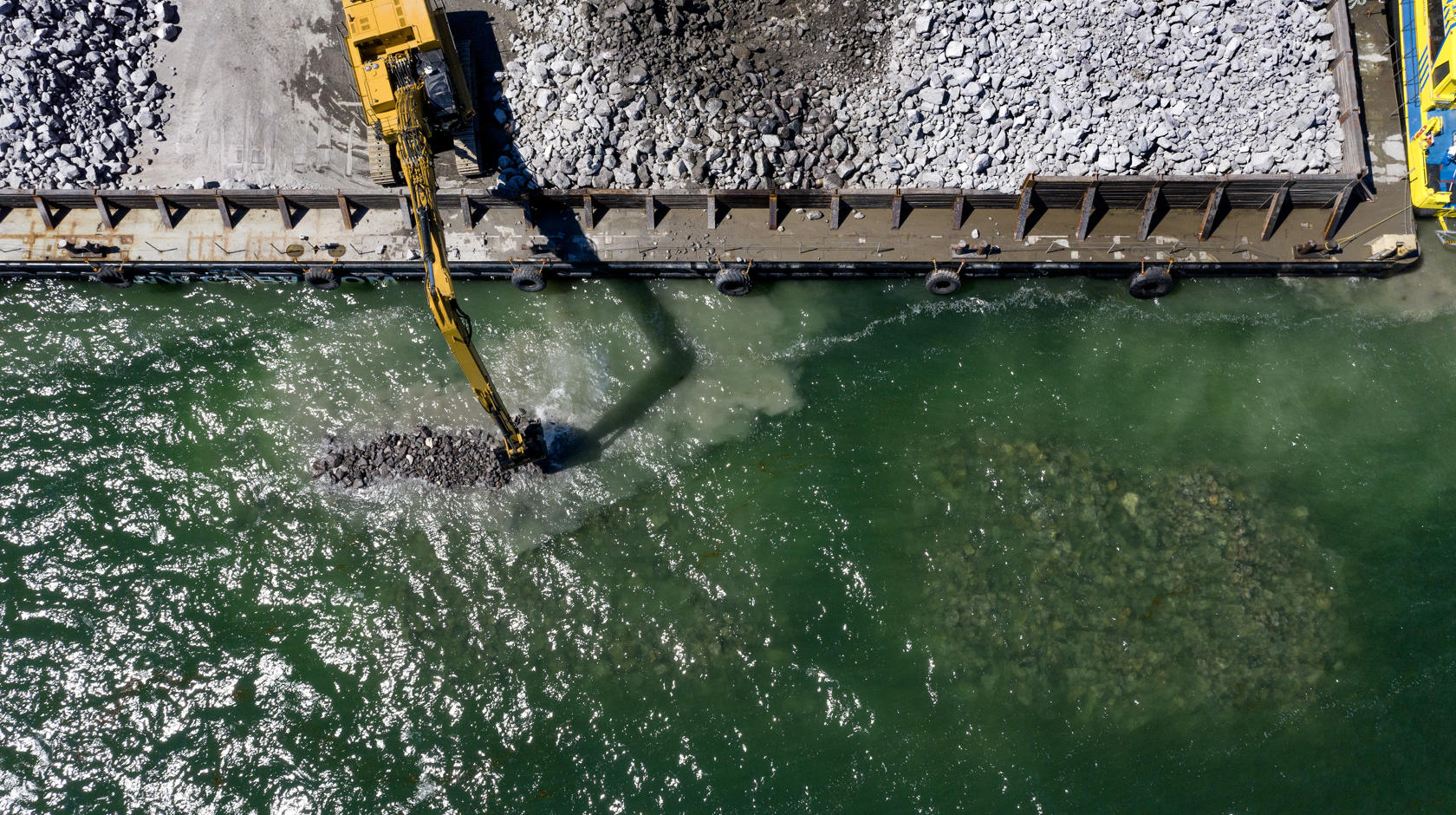 English: An excavators drops boulders into the sea to construct breakwaters.//Dansk: En gravemaskin lægger kampestenene i havet for at bygge bølgebryderne.//Deutsch: Ein Bagger versenkt Granitbrocken im Meer, um Wellenbrecher zu errichten.
