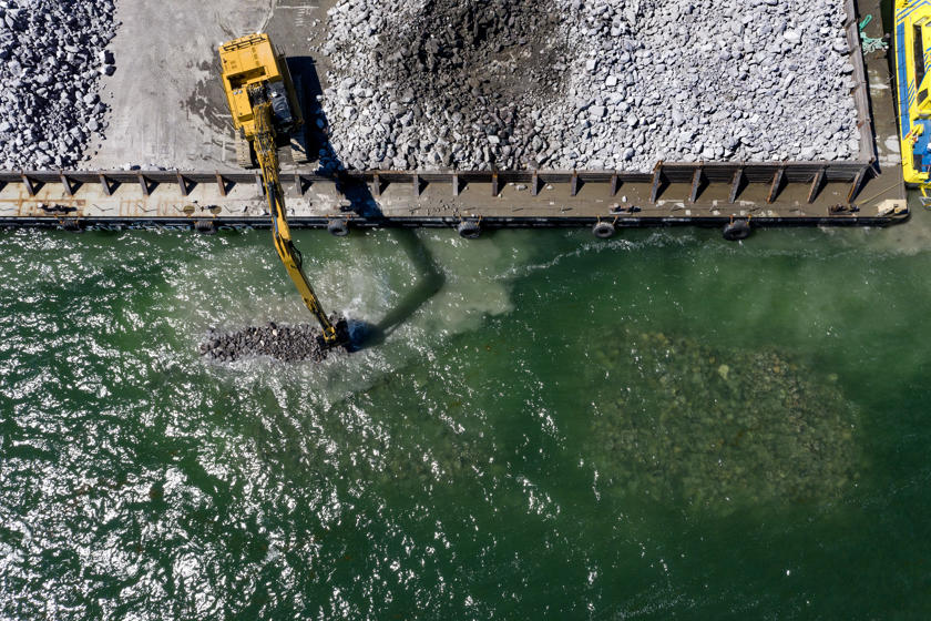 English: An excavators drops boulders into the sea to construct breakwaters.//Dansk: En gravemaskin lægger kampestenene i havet for at bygge bølgebryderne.//Deutsch: Ein Bagger versenkt Granitbrocken im Meer, um Wellenbrecher zu errichten.