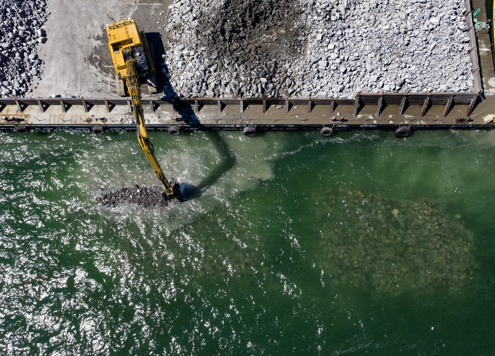 English: An excavators drops boulders into the sea to construct breakwaters.//Dansk: En gravemaskin lægger kampestenene i havet for at bygge bølgebryderne.//Deutsch: Ein Bagger versenkt Granitbrocken im Meer, um Wellenbrecher zu errichten.