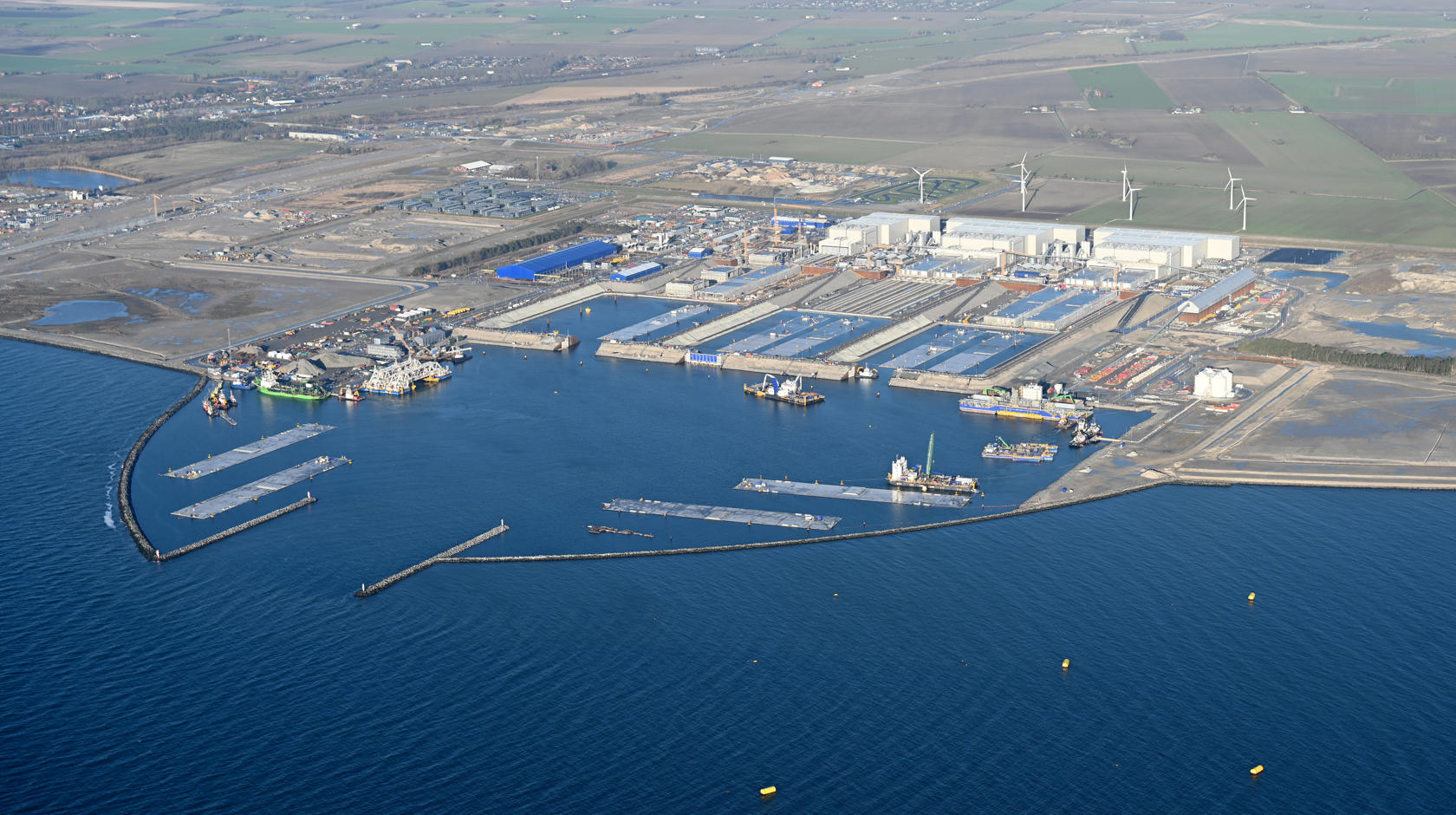 Den danske tunnelbyggeplads. I arbejdshavnen ligger arbejdsskibe og tunnelelementer. Man kan se de tre store hvide haller, hvor tunnelelementerne produceres.