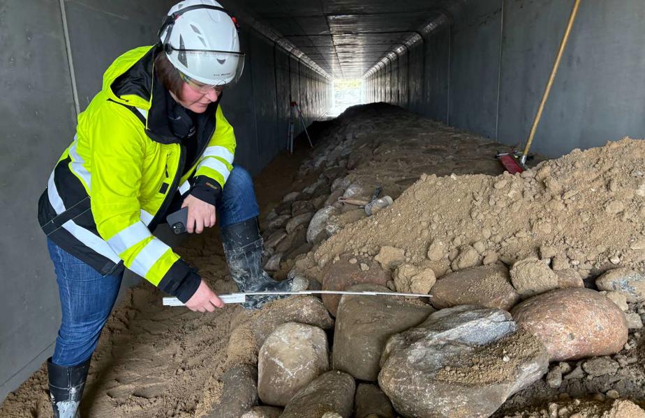 Eine Mitarbeiterin in gelber Jacke misst mit einem Zollstock Steine in dem Wildtierdurchlass. Das ist eine lange Röhre aus Beton.