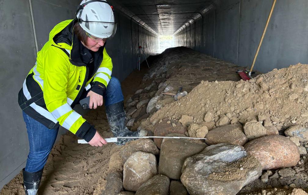 Eine Mitarbeiterin in gelber Jacke misst mit einem Zollstock Steine in dem Wildtierdurchlass. Das ist eine lange Röhre aus Beton.